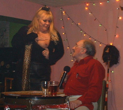 Woman with leopard skin scarf exposes one bussem to a shocked looking Gordon Cragg, who is seated at the bingo table, an array of fairy lights twinkle gently in the back ground conjuring the image of Blackpool illminations.